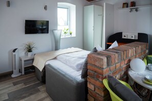 Een slaapkamer met een bed, een televisie, een raam met planten, een bakstenen muur en een klein tafeltje met een kop.