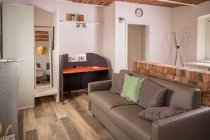 A room with a sofa, a desk, a brick accent wall, a clock, and a door leading to another room.