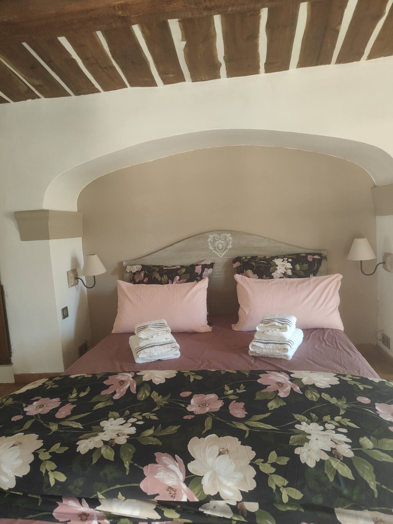 Une chambre à coucher avec un lit, une couvre-lit à motifs floraux, deux oreillers, deux serviettes pliées et une tête de lit ornée d’un motif en forme de cœur.