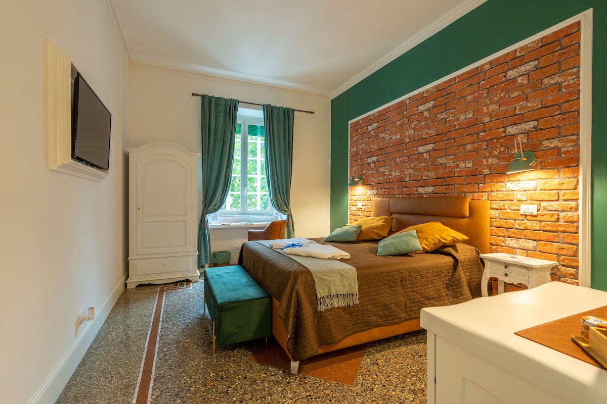 A bedroom with a brick wall, a bed with pillows, a bench, a window with curtains, and a TV mounted on the wall.