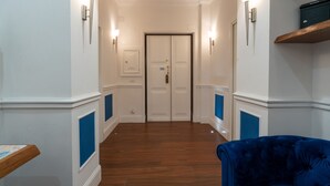 Un couloir avec un plancher en bois, des murs blancs et des touches de bleu. On aperçoit une porte au fond et une chaise bleue au premier plan.