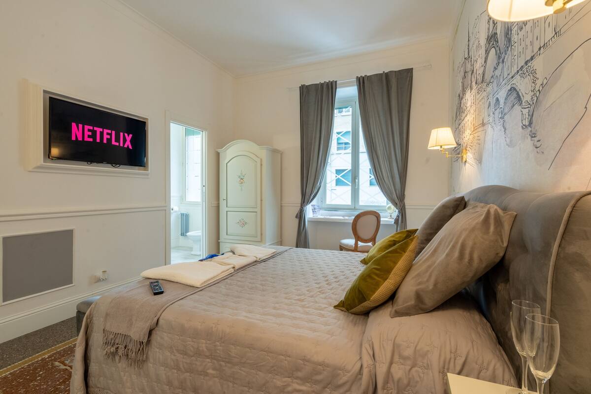 A hotel room with a bed, a TV displaying 'NETFLIX', a wardrobe, a window with curtains, and a lamp.