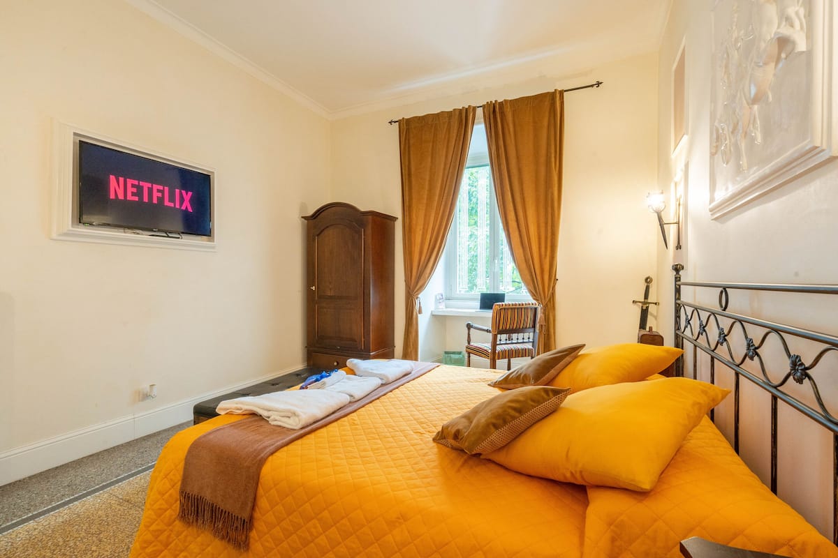 A hotel room with a bed, a TV displaying 'NETFLIX', a window with curtains, a wooden wardrobe, and a chair.