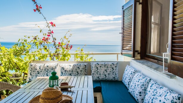 Un balcon avec une table en bois, un chapeau en fibres tressées et une lanterne verte. On y trouve un canapé avec des coussins à motifs floraux et une vue sur la mer.