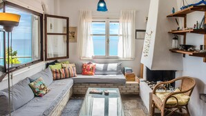 Un salon chaleureux avec un canapé, une table basse en verre et un fauteuil en osier. Une fenêtre, ornée de rideaux, offre une vue sur la mer.