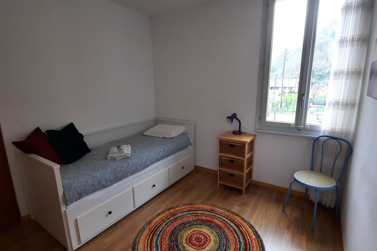 Habitación pequeña con cama individual, suelo de madera, ventana con cortinas, mesita de noche de madera y una silla.