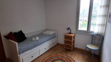 Habitación pequeña con cama individual, suelo de madera, ventana con cortinas, mesita de noche de madera y una silla.