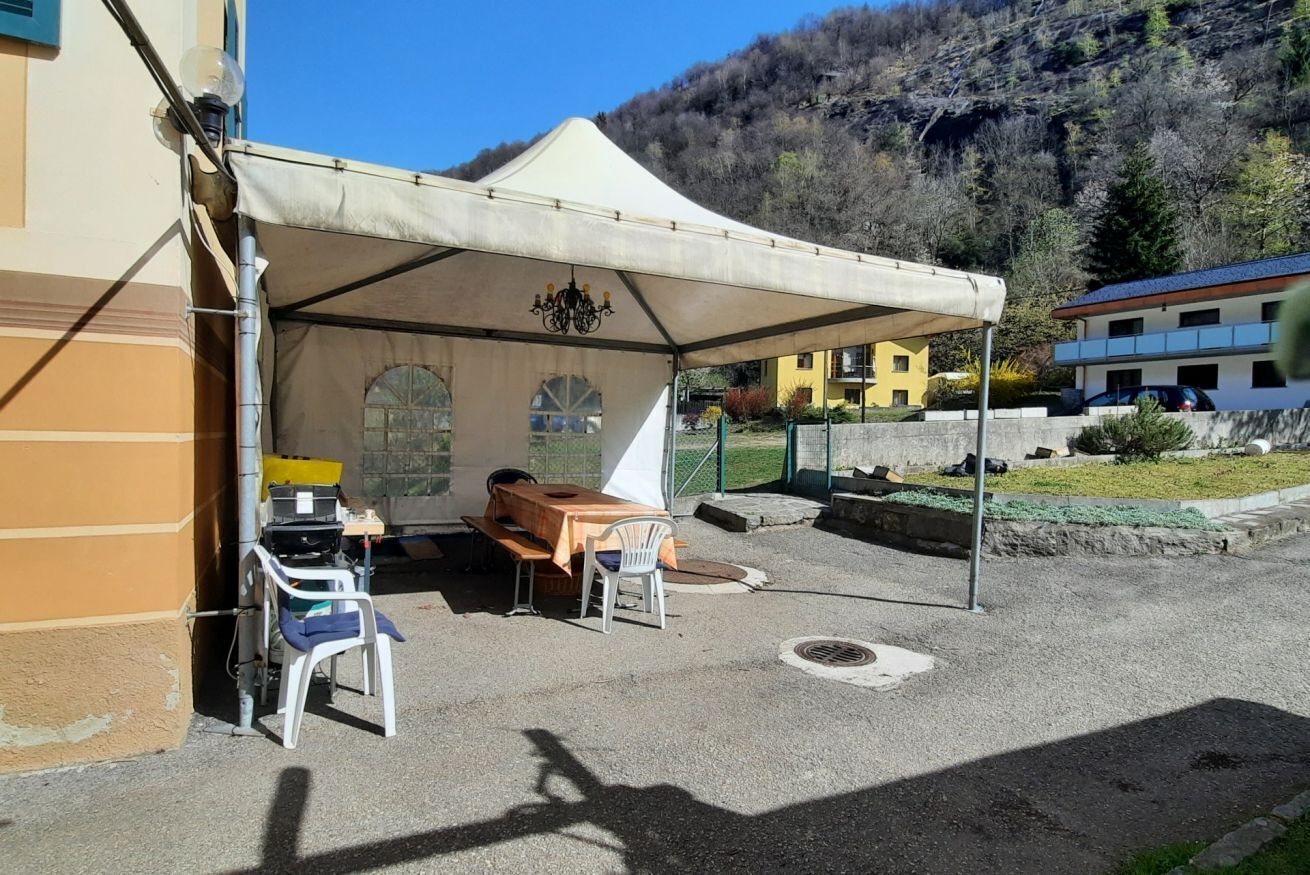 Ein Außenbereich mit Tisch und Stühlen unter einem Zelt, ein Gebäude mit gelber Fassade und eine gebirgige Landschaft im Hintergrund.