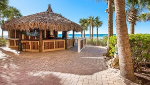 A beachside bar with a thatched roof, surrounded by palm trees and a paved walkway.