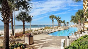 A pool area with lounge chairs, palm trees, and a view of the beach and pier.