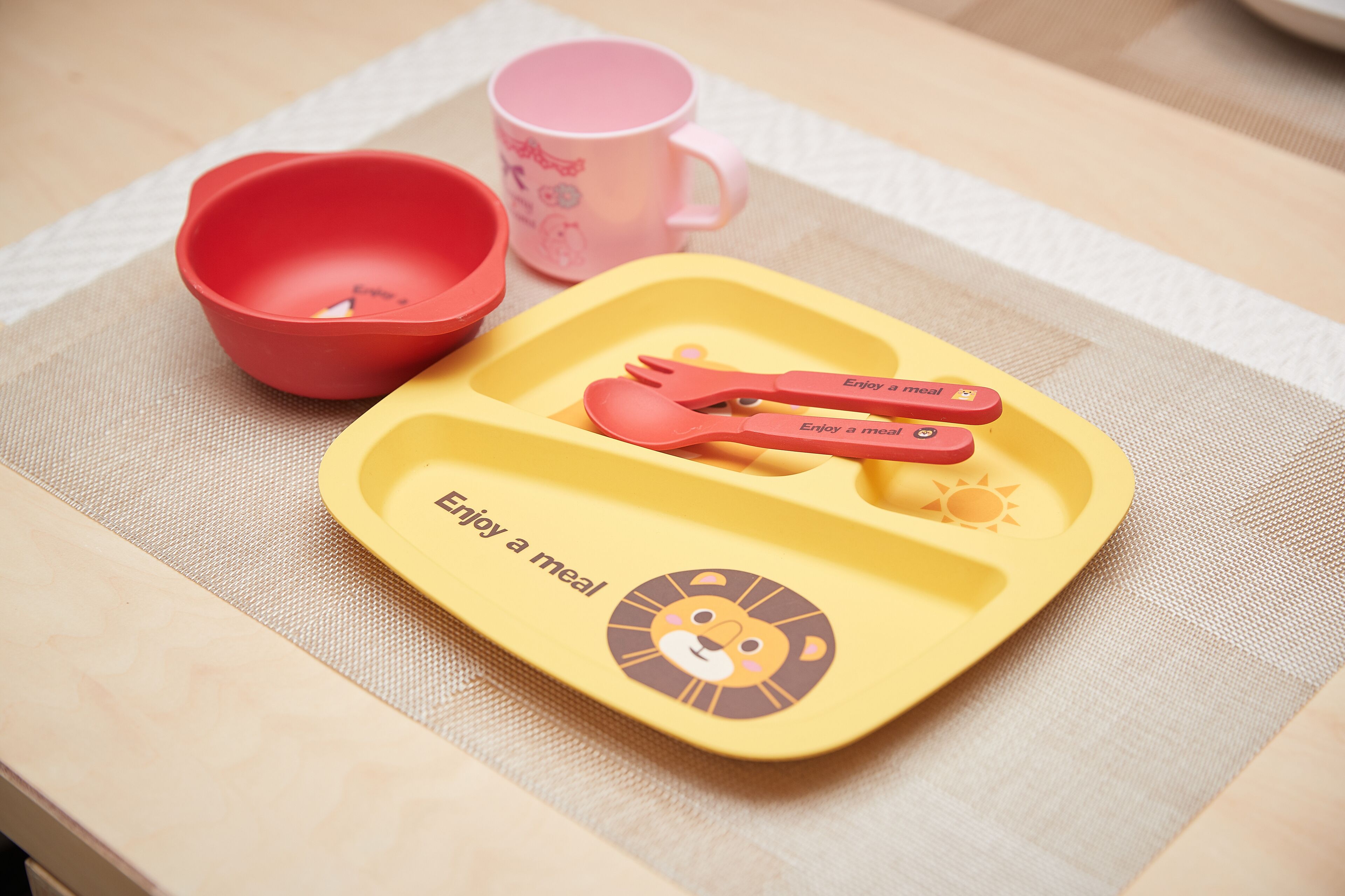 A tray with a lion face and the text "Enjoy a meal" on a table with a pink mug and a red bowl.