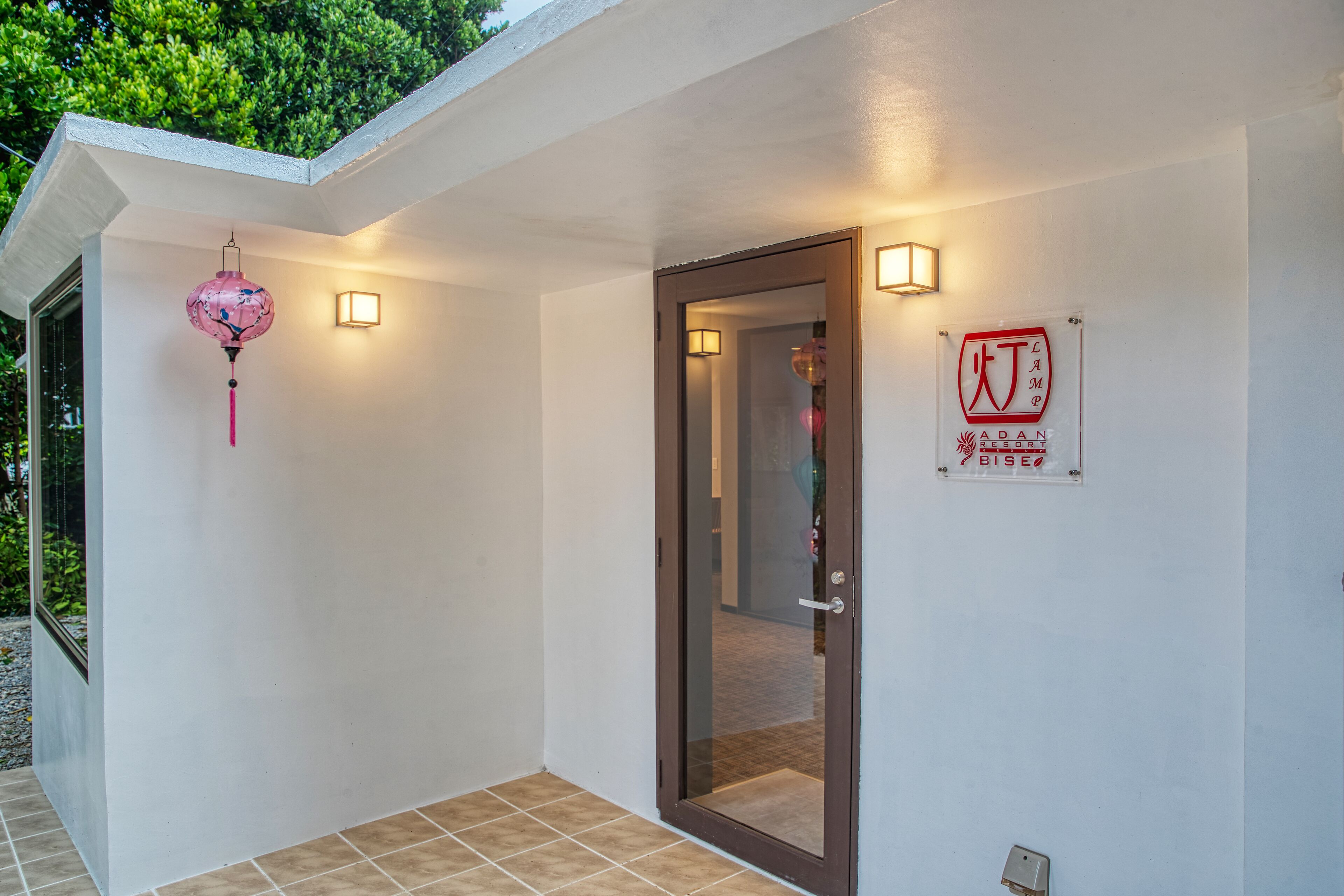 A hotel entrance with a glass door, a sign indicating 'ADAN CAMP BISEO', and a pink lantern hanging on the wall.