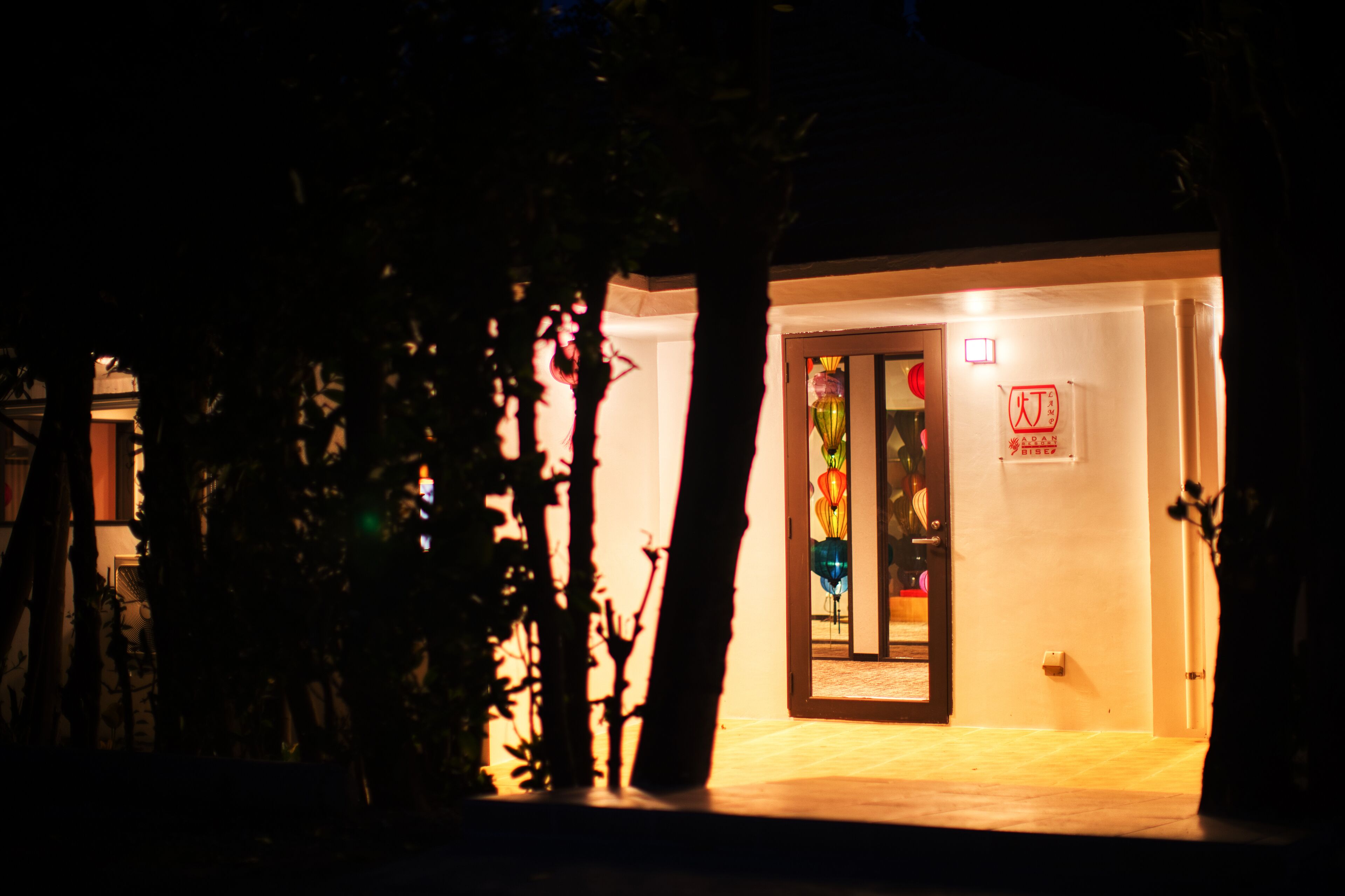 A building with a lit entrance, a door with a sign, and a small garden area with trees.