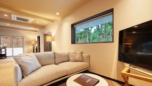 A modern hotel room with a beige sofa, a round coffee table, a flat-screen TV, and a window with a view of greenery. - ADAN RESORT Lamp (Motobu)