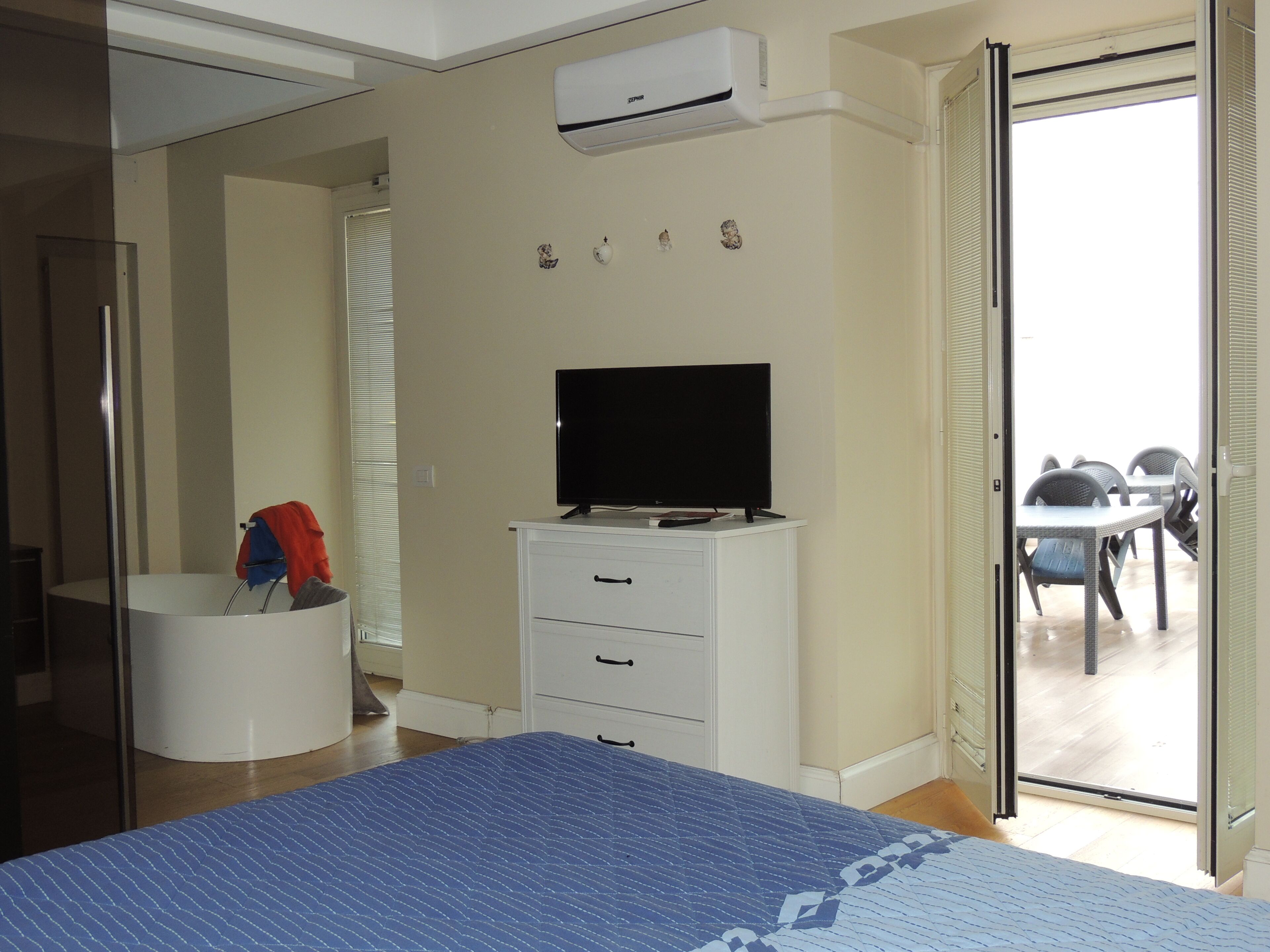 Ein Schlafzimmer mit Bett, Fernseher auf einer Kommode, ein Badezimmer mit Dusche und ein Balkon mit Sitzmöbeln.