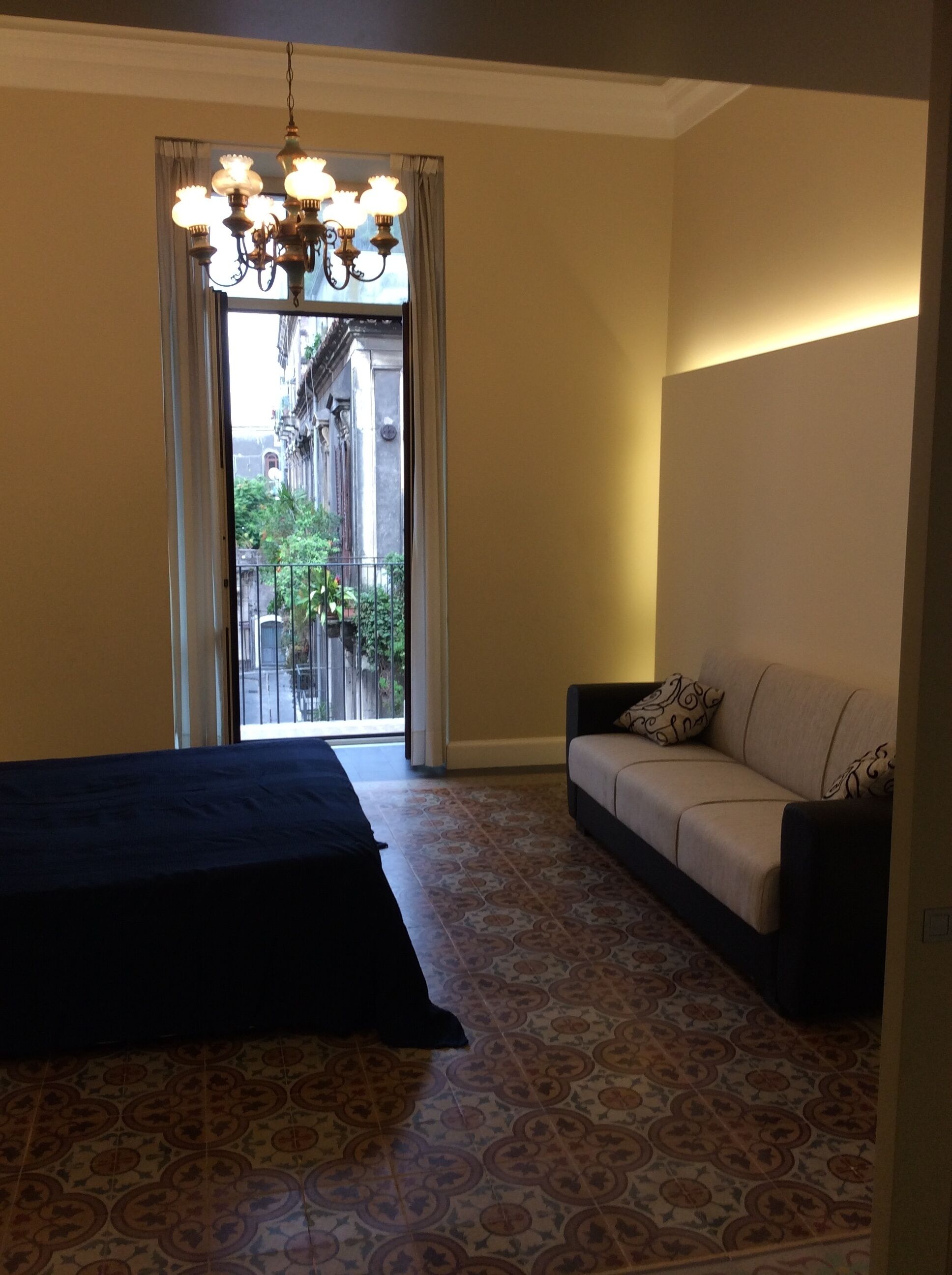 Ein Hotelzimmer mit Bett, Sofa, einem gemusterten Boden, einer Chandelierleuchtre und Blick auf Gebäude durch ein Fenster.
