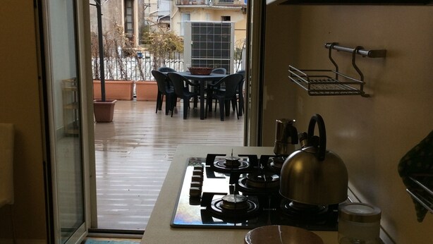 A compact kitchen with a gas stove, kettle, and two glasses on the counter. A sliding glass door leads to an outdoor patio with a table and chairs. - Chicca suite with terrace in the heart of the historic center of Catania (catania)