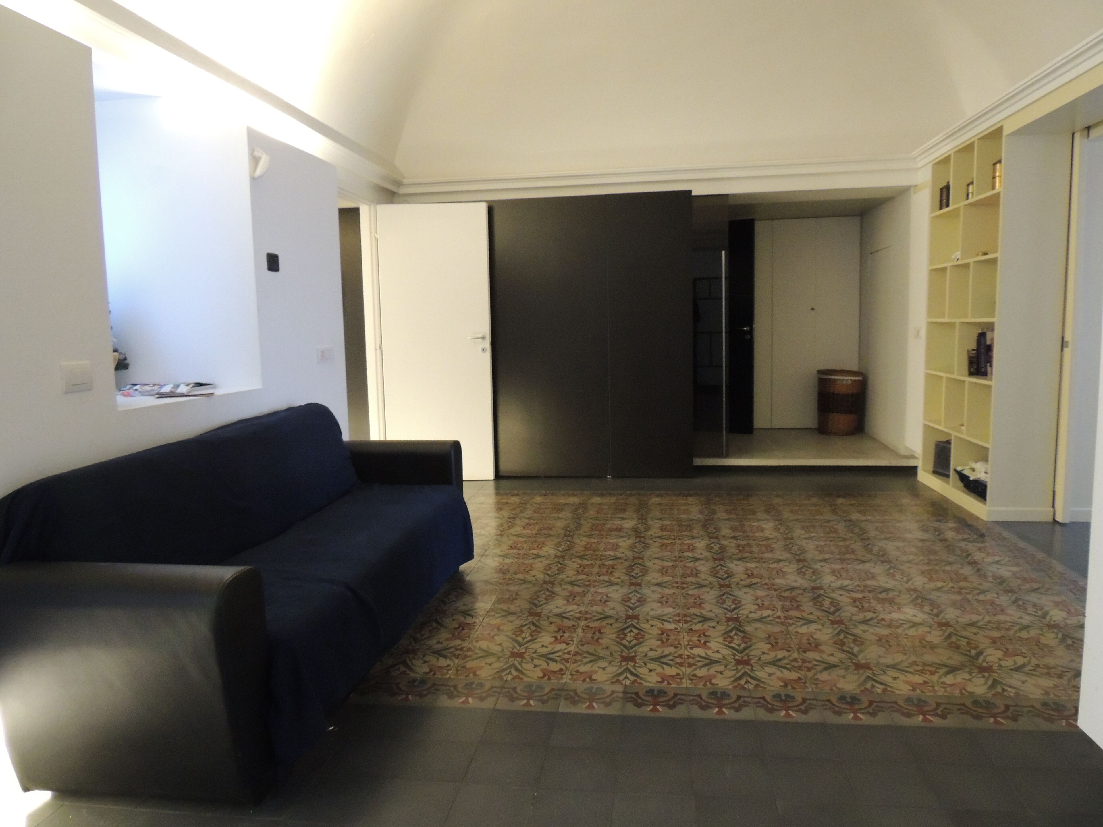 A living room with a dark blue sofa, a patterned rug, and a bookshelf with various items.