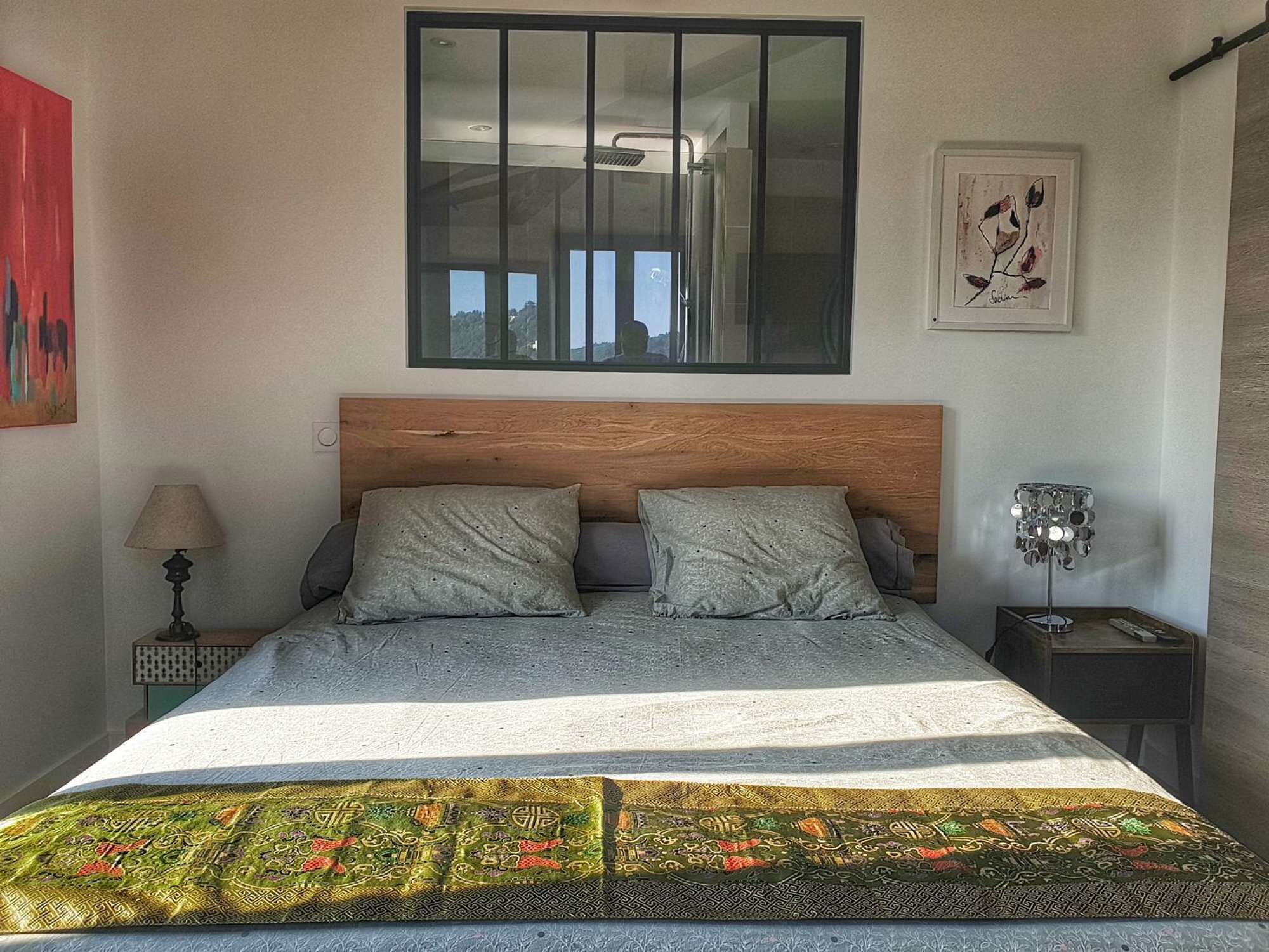 Un dormitorio con cabecera de madera, dos almohadas, colcha estampada, mesita de noche con lámpara y una obra de arte enmarcada en la pared.