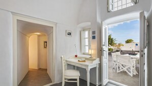 Un escritorio blanco con una silla, una mesita con una copa de vino, y vista a un patio exterior con mobiliario blanco.