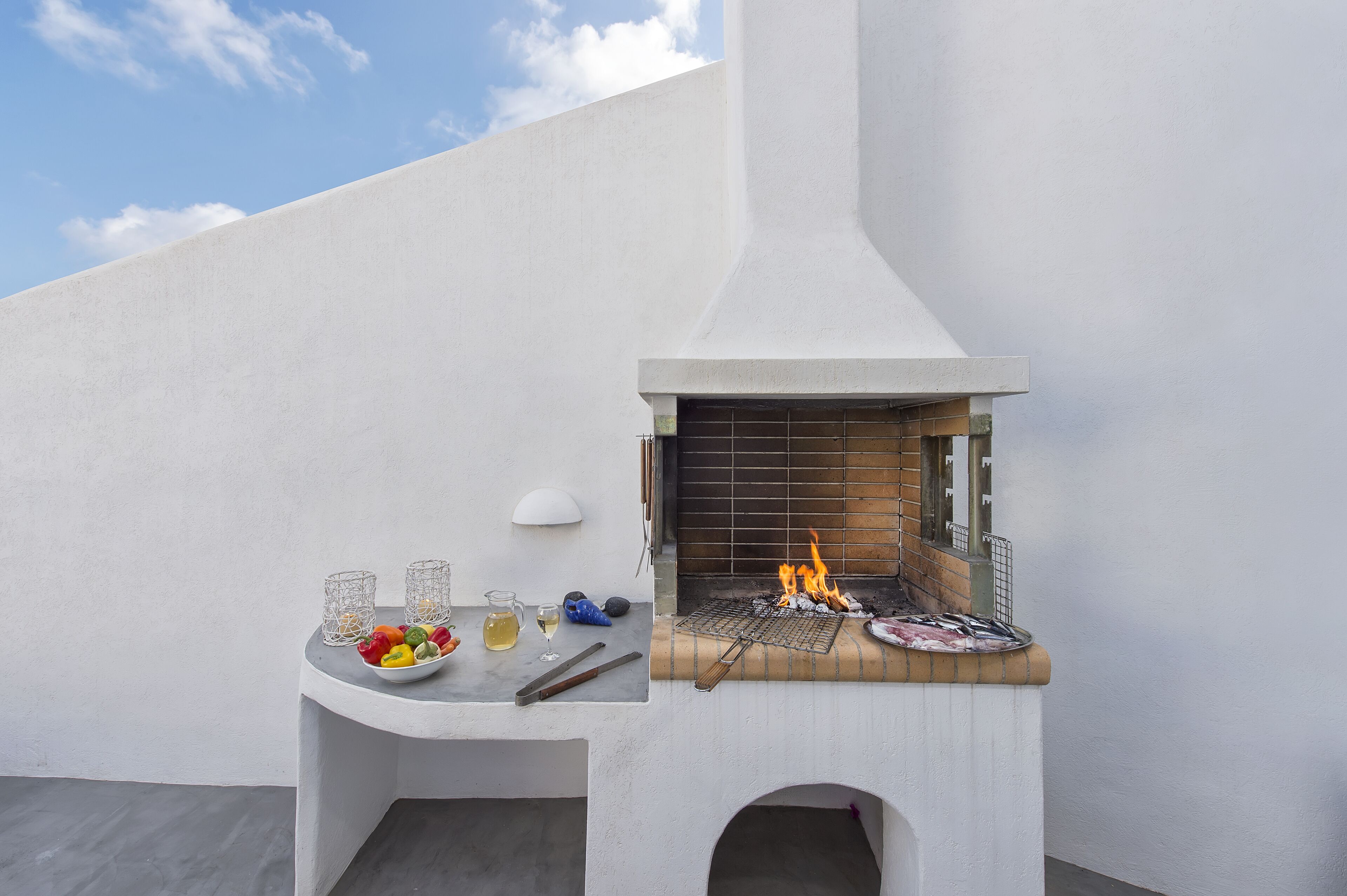 A modern outdoor grill area with a white wall, a fire burning, and a table with food and drinks.