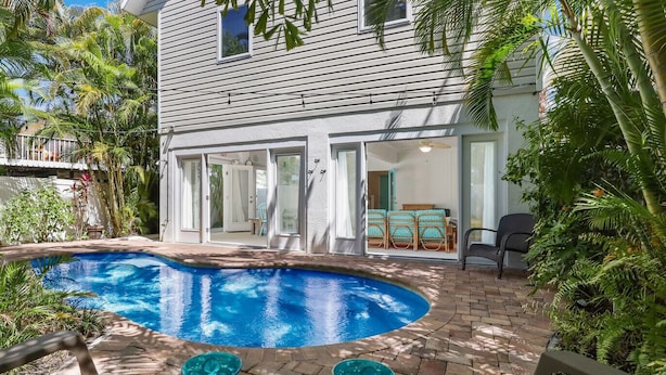 A swimming pool with a curved edge, surrounded by a brick patio and lush greenery. - Wish Upon a Starfish by Beach Boutique Rentals (Holmes Beach)