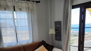 Ein Wohnzimmer mit braunem Sofa, einem kleinen Tisch mit Topfpflanze und Blick auf das Meer durch eine Glastür.