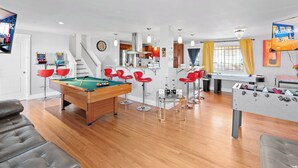 Een moderne woonkamer met een pooltafel, rode stoelen, een tafelvoetbaltafel en een keuken op de achtergrond.