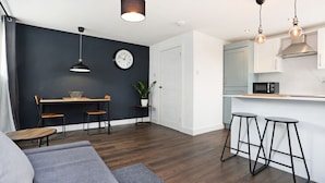 Een moderne keuken met een magnetron, een klok en een plant. Er is een eethoek met een tafel en stoelen, en een bar met krukken.