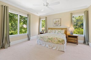 Une chambre à coucher avec un grand lit, une table de chevet, un ventilateur de plafond et des fenêtres avec des rideaux.