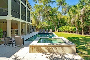 Une piscine avec des chaises longues, un patio couvert et une grande maison dotée de nombreuses fenêtres.