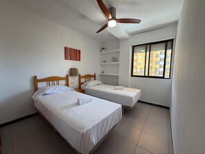 A room with two beds, a ceiling fan, a window with a view of buildings, and a shelf with items.