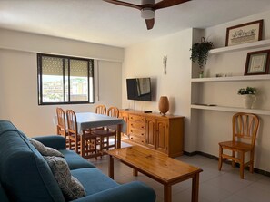Ein Wohnzimmer mit einer Couch, einem Holzesstisch und Stühlen. An der Wand ist ein Fernseher, ein Deckenventilator und ein Fenster mit Jalousien.