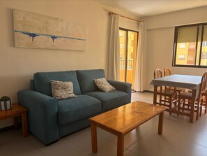 Ein Wohnzimmer mit einer Couch, einem Holztisch, einem Esstisch mit Stühlen, einem Fenster mit Vorhängen und einem Gemälde an der Wand.