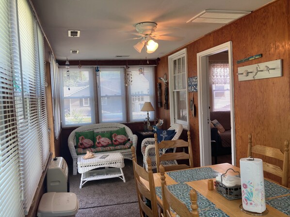 A cozy room with a wooden wall, a ceiling fan, a wicker sofa, a dining table with a patterned table runner, and a wall-mounted sign.