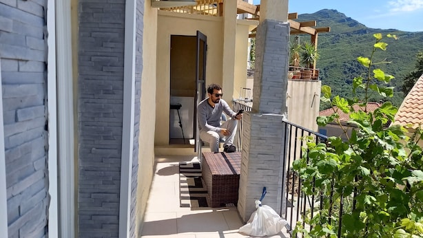 Un homme est assis sur un balcon, avec une vue sur les montagnes, un panier en osier et un sac en plastique blanc.