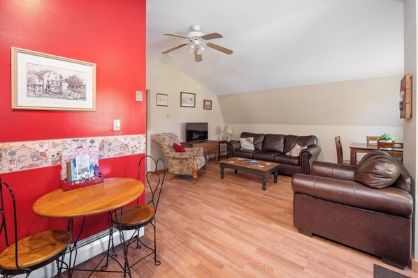 Una sala de estar con pared roja, ventilador de techo, chimenea, sofá de cuero marrón, mesa de centro de madera y un comedor con mesa y sillas.
