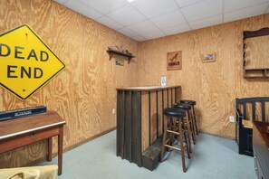 A room with a wooden wall, a 'DEAD END' sign, a bar area with stools, a table with a box labeled 'PICTIONARY', and a chair.