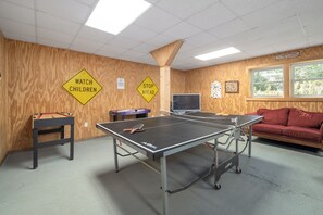 A room with ping pong tables, a TV, and a red couch. There are warning signs on the wall and a window with a view.