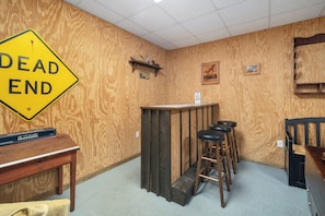 A room with a wooden wall, a 'DEAD END' sign, a bar area with stools, a table with a box labeled 'PICTIONARY', and a chair.
