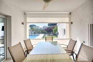 A conference room with a large table and six chairs, large windows with a view of a swimming pool and greenery, and a ceiling fan. - FALCONERA VILLA CANAL  3 HA ,PISCINA,AMARRE,WIFI,PARKING (Empuriabrava)