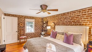 Une chambre avec un mur en briques, un ventilateur de plafond, un lit avec une literie à motifs et une chaise avec un coussin.