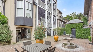 Une cour avec une terrasse en briques, des sièges extérieurs, une fontaine et un bâtiment doté d’un balcon et d’un mur couvert de lierre.