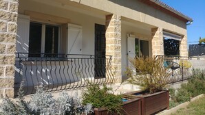 Une maison à deux étages avec une façade en pierre, un toit en tuiles et un porche couvert doté d’une rambarde en métal.