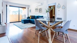 Un salon moderne avec une table à manger en verre, des chaises grises, un canapé bleu et une grande fenêtre.