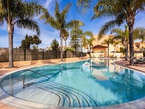 A swimming pool with lounge chairs and palm trees, surrounded by a fence and buildings.