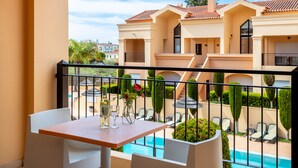 A balcony with a table, two wine glasses, and a bottle. In the background, there is a swimming pool and a building with a red-tiled roof.