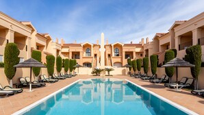 A pool area with lounge chairs and umbrellas, surrounded by buildings with a symmetrical architectural design.