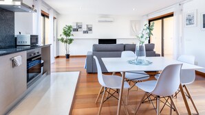 A modern kitchen with built-in appliances, a dining area with a white table and chairs, a grey sofa, and a hanging light fixture.
