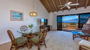 A dining area with a glass table and wicker chairs, a ceiling fan, a TV, and a view of a mountainous landscape through large sliding doors. - Maui Kamaole #L-203 Full Ocean View, A/C Throughout, Wifi, Great Location (Kihei)